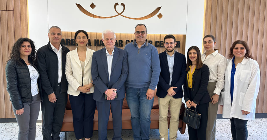 Group of nine people standing indoors in front of a sign that reads 'Barbara Najar Cancer Center', posing for a photo with a wooden paneled wall and beige curtains in the background