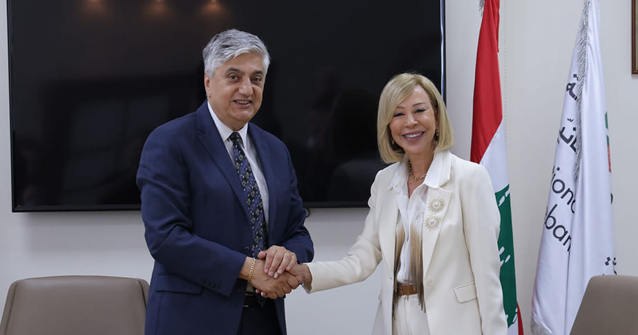LAU President Chaouki T. Abdallah shaking hands with Lebanon's First Lady Nehmat Aoun in an office setting with the LAU and Lebanese flags in the background.