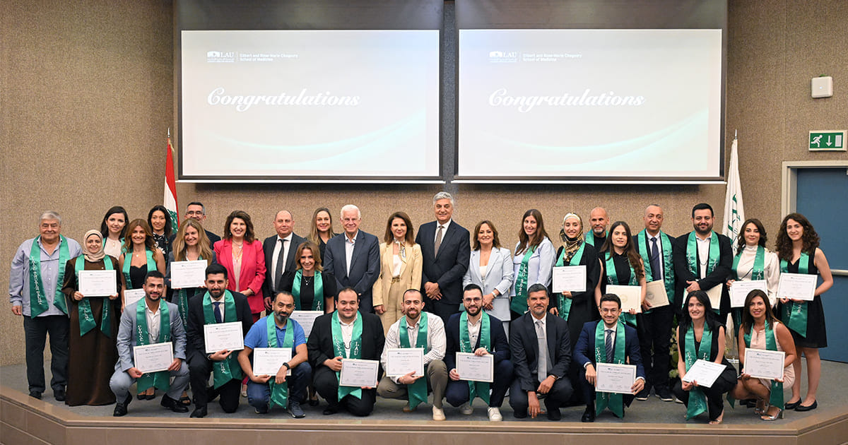 Group photo of graduates od the Clinical Simulation Diploma and faculty members posing on stage with certificates and green stoles in front of two screens displaying the word "Congratulations."