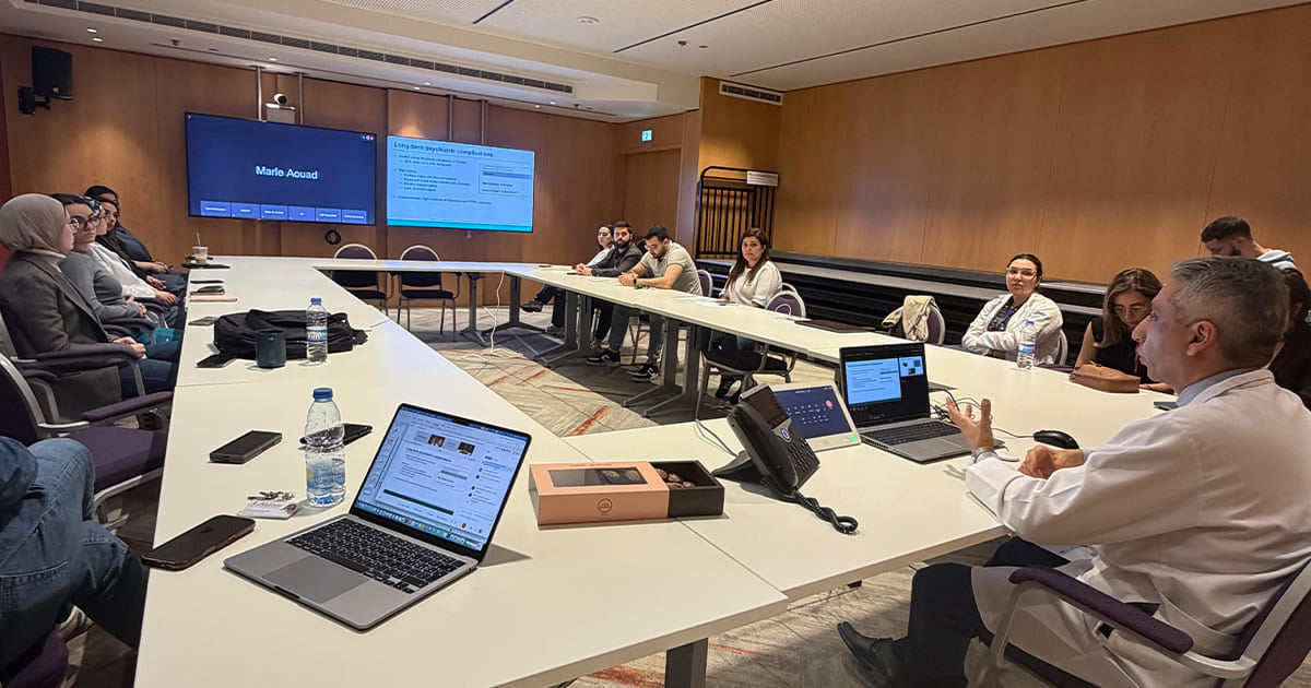 Conference room with people sitting around U-shaped tables, some with laptops open, engaging in a meeting or presentation with two large screens on the wall displaying information.