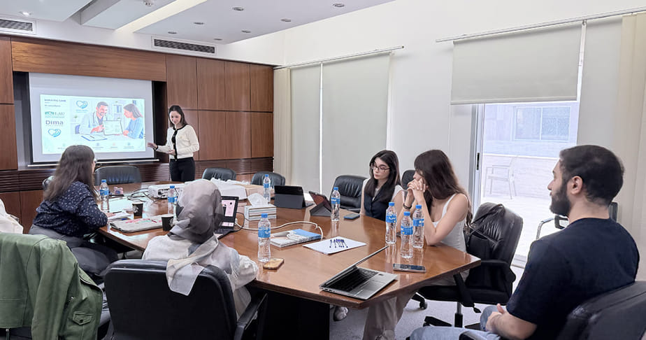 Meeting room with seven people attending a presentation, one person standing and pointing at a screen displaying a presentation slide, others seated around a table with laptops and notebooks.