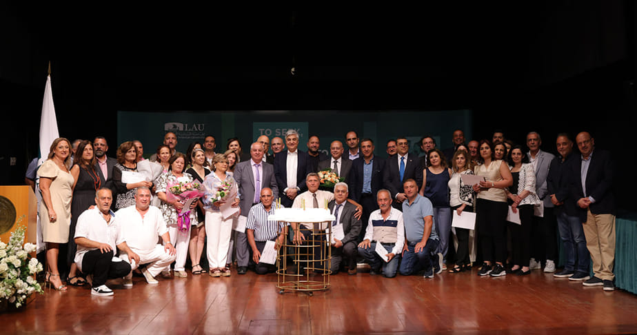 LAU's longest serving staff gathered on a stage for a formal event, some holding bouquets and certificates, with a table and a cake in front of them.