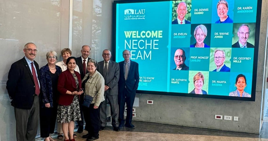 Group of professionals standing indoors next to a digital screen welcoming the NECHE team at Lebanese American University