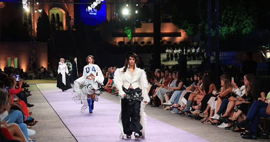 Fashion models walking on an outdoor runway at night with an audience seated on both sides watching the show.