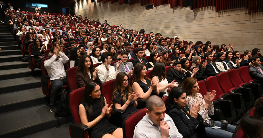 Audience in formal attire sitting in a theater-style auditorium clapping.