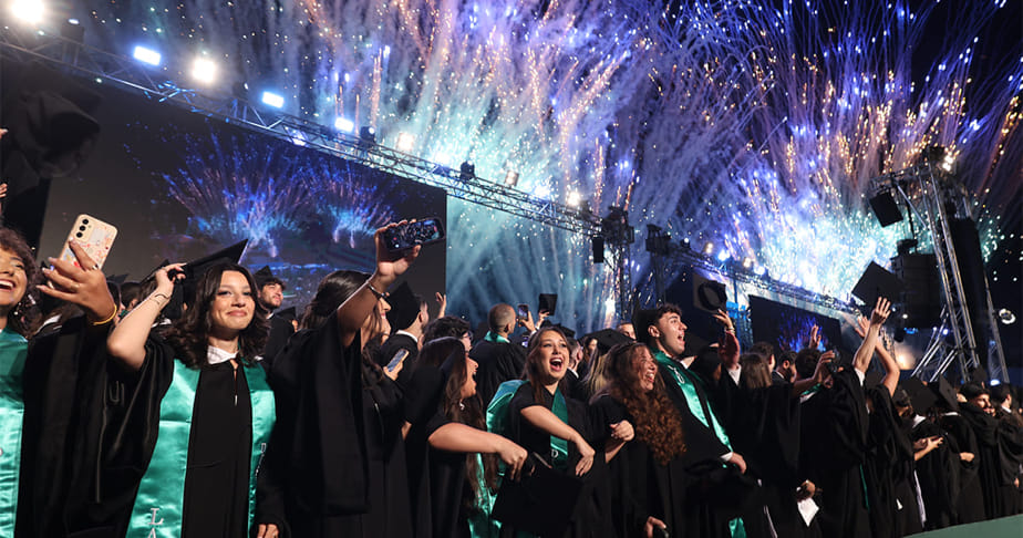 Graduates in black gowns and teal stoles celebrating with fireworks at a nighttime outdoor graduation ceremony.
