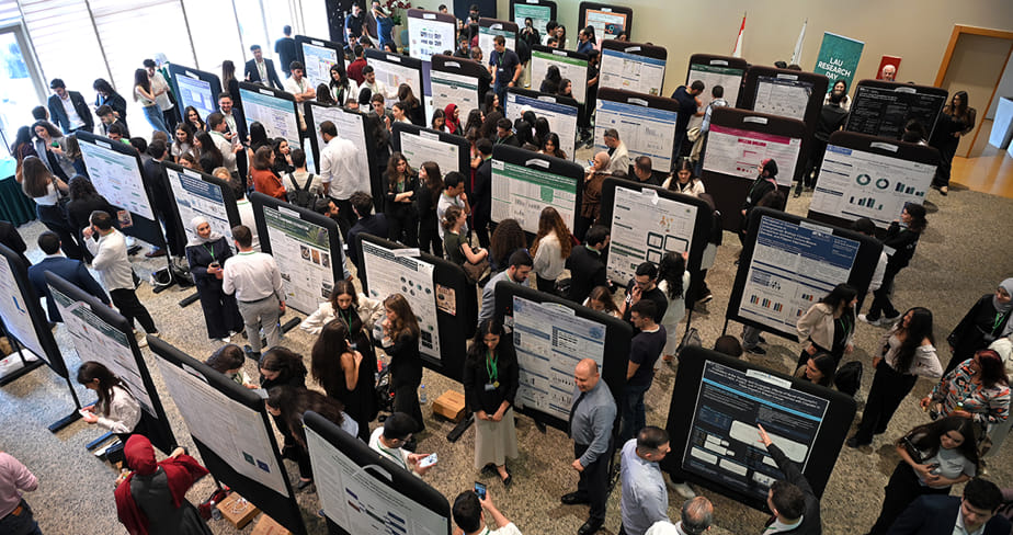 Poster presentation event with numerous people discussing and viewing scientific or academic posters displayed on black boards in a large indoor space.