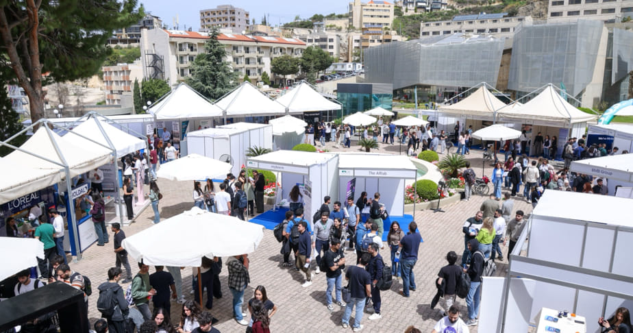 Job fair with multiple white exhibition booths and tents, numerous people walking and interacting, and buildings in the background.