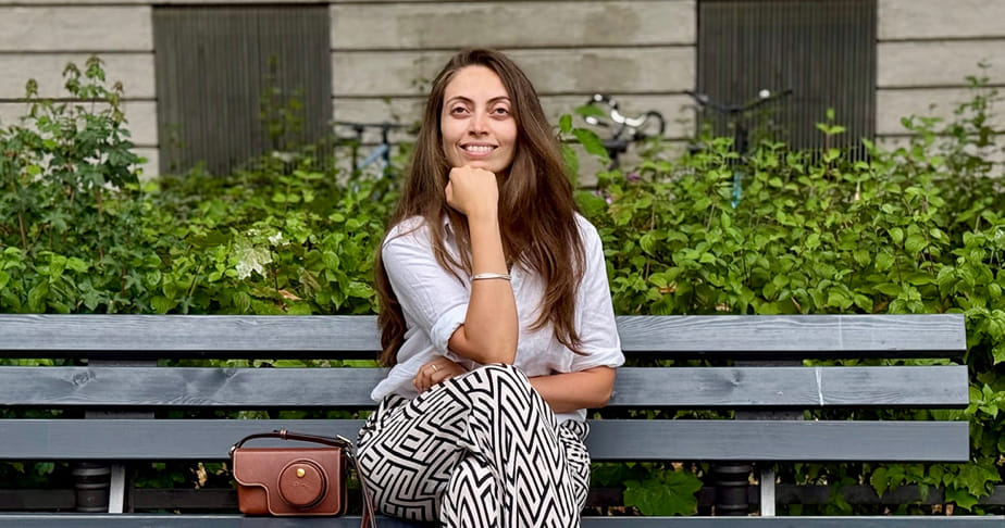 Graphic design alumna Patricia Farah (BS’12) sitting on a gray bench outdoors with green bushes in the background.