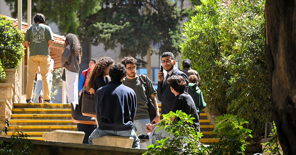 Group of students socializing outdoors near a staircase surrounded by greenery on a sunny day in Beirut campus.
