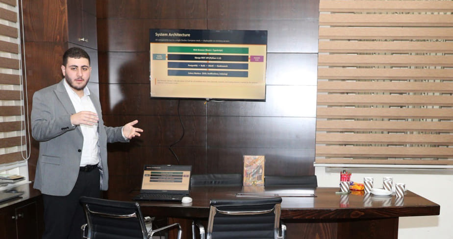 A computer science student giving a presentation in a conference room with a laptop and a screen displaying system architecture.